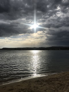 Sun shining through dark clouds over a calm bay with light reflecting on water and sandy beach in foreground