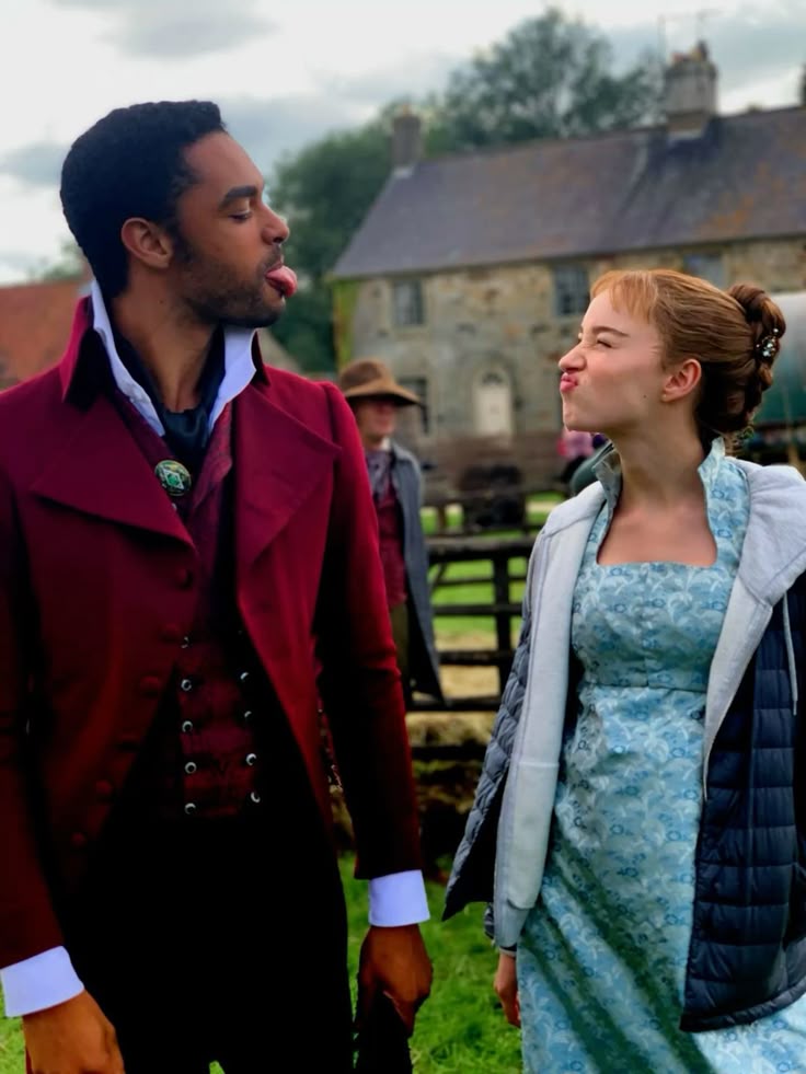 Man in red Regency coat and woman in blue gown looking at each other outdoors near stone buildings