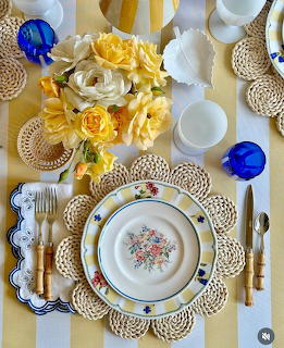 Table setting with floral plates, woven placemats, yellow roses, gold cutlery and blue glassware