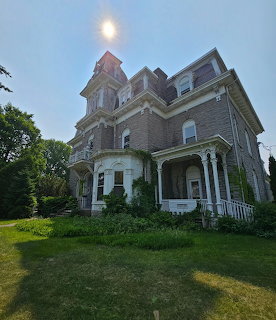 Gray Victorian mansion with square tower and front porch surrounded by overgrown grass and trees