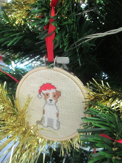 Cross-stitch Christmas ornament of Jack Russell dog in Santa hat with gold tinsel border hanging on tree