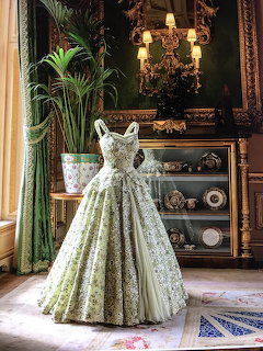 White floral Victorian ball gown on display stand in ornate period room with gilt mirror and green drapes