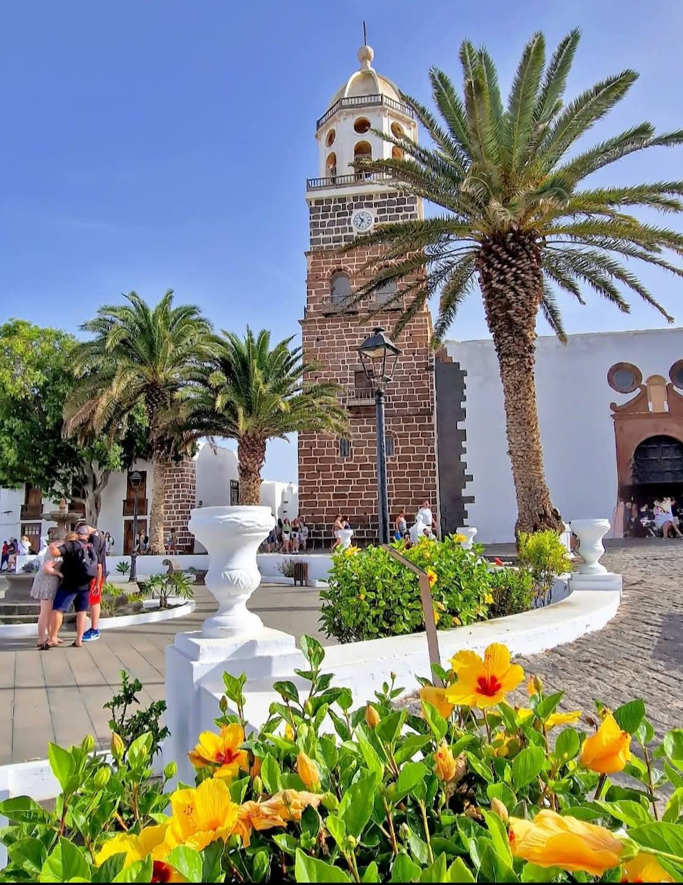 Lanzarote plaza with stone church bell tower, palm trees, white railings, and orange hibiscus flowers under blue sky