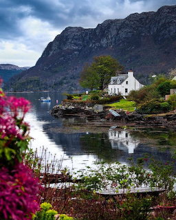 White cottage on a rocky lakeshore with mountain backdrop, calm water reflections, and pink flowers in front