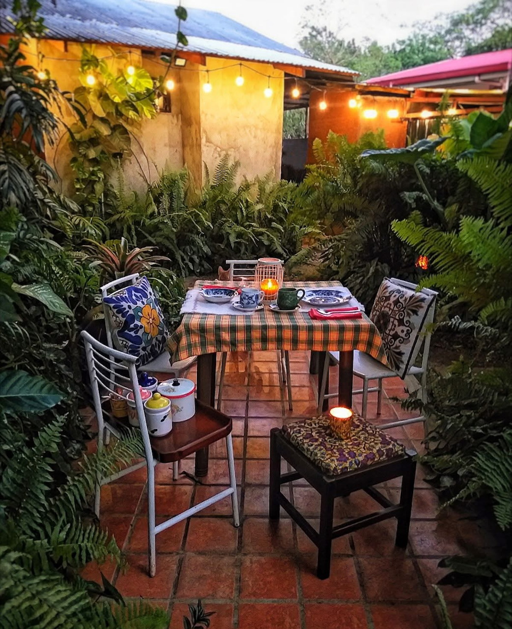 Outdoor patio dining table with checkered cloth surrounded by dense green plants and warm string lights at night.