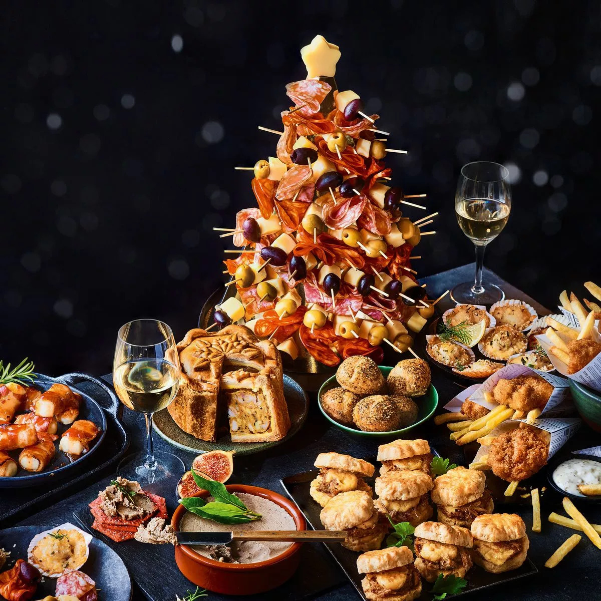 Festive appetizer spread with bacon tower centerpiece, assorted finger foods, and wine glasses on dark background