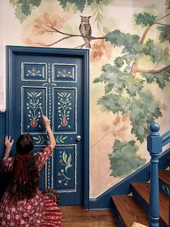 Woman reaching toward an ornately painted blue door beside a wall mural with trees and an owl