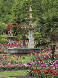 Multi-tiered stone fountain in formal garden with vibrant pink, purple, and red flowers and palm trees