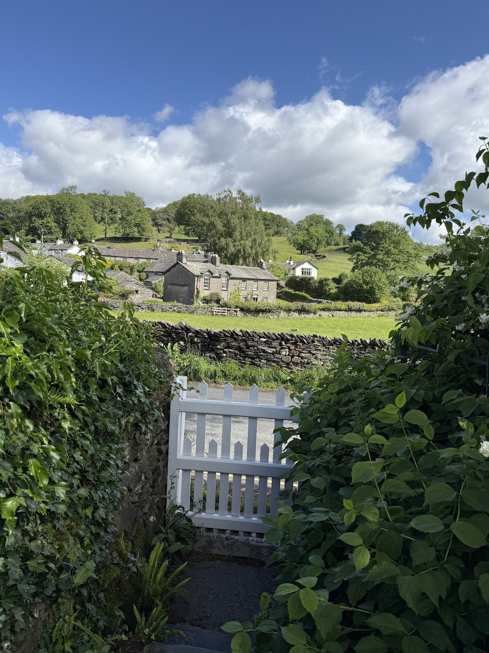White garden gate between hedges, stone cottages and green hills under blue sky beyond