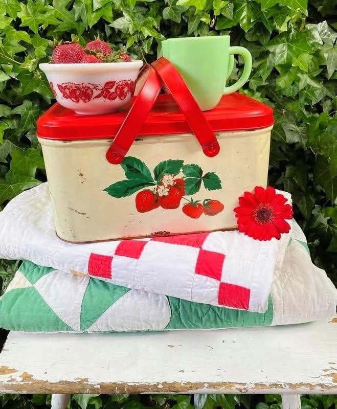 Vintage strawberry breadbox with green mug, berry bowl, red flower, and folded quilts against ivy