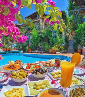 Breakfast table with various dishes and orange juice by a pool with pink flowers and greenery