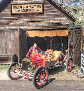 Red 1912 Ford Speedster vintage car in front of old wooden blacksmith shop with man in period clothing
