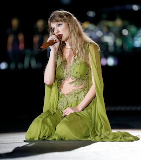 Woman in green lace outfit and cape singing into microphone while kneeling on stage
