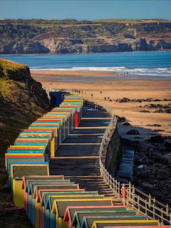 Rows of colorful beach huts along a coastal walkway with sandy beach, cliffs, and ocean beyond