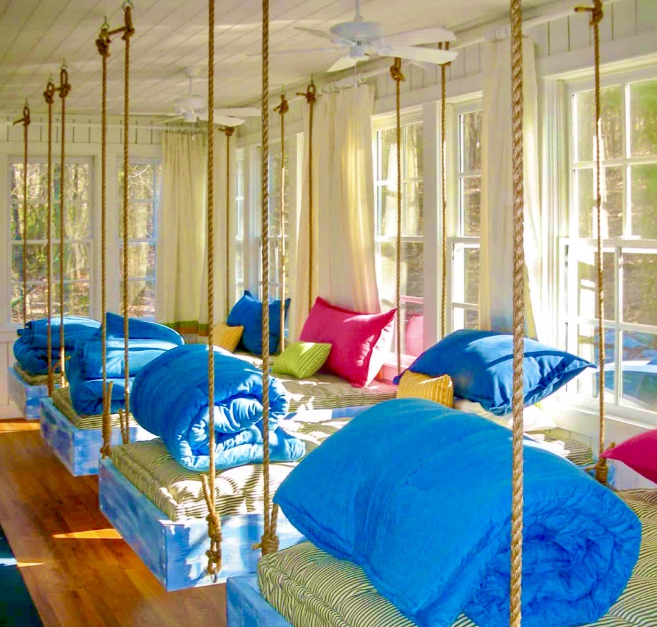 Sunlit sleeping porch with rope-hung beds, blue pillows, and tall windows