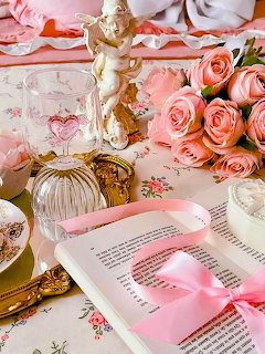 Pink aesthetic flatlay with roses, open book, cherub figurine, and vintage glass items on floral fabric