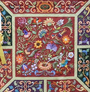 Ornate quilt with embroidered flowers and scrollwork on red fabric with decorative border