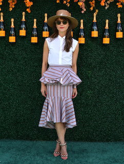 Woman in sunglasses and hat standing by a green hedge wall with bottles above