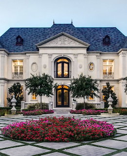White mansion with arched entrance, lit windows, and colorful flower bed in front courtyard at twilight