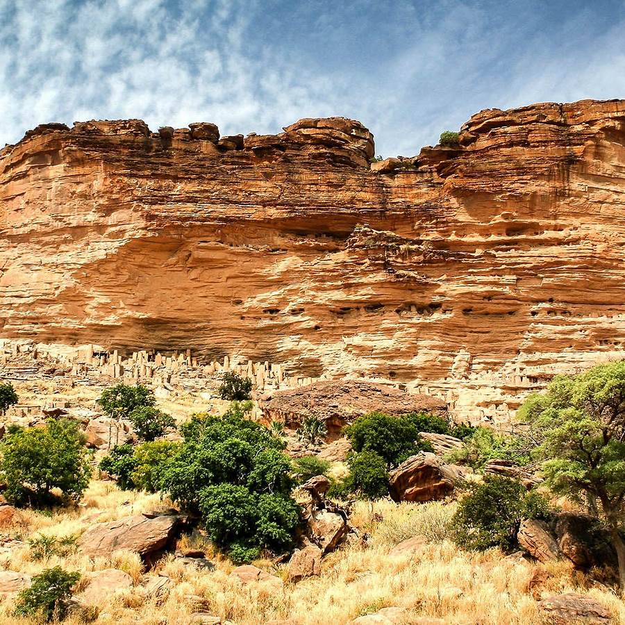 Sandstone cliff above dry grass and green shrubs beneath a lightly clouded sky