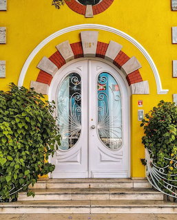 Ornate white arched double door with stained glass panels in a bright yellow wall with green plants