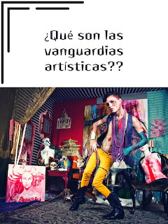Tattooed artist in yellow pants posing in a colorful, cluttered art studio with paintings and sculptures