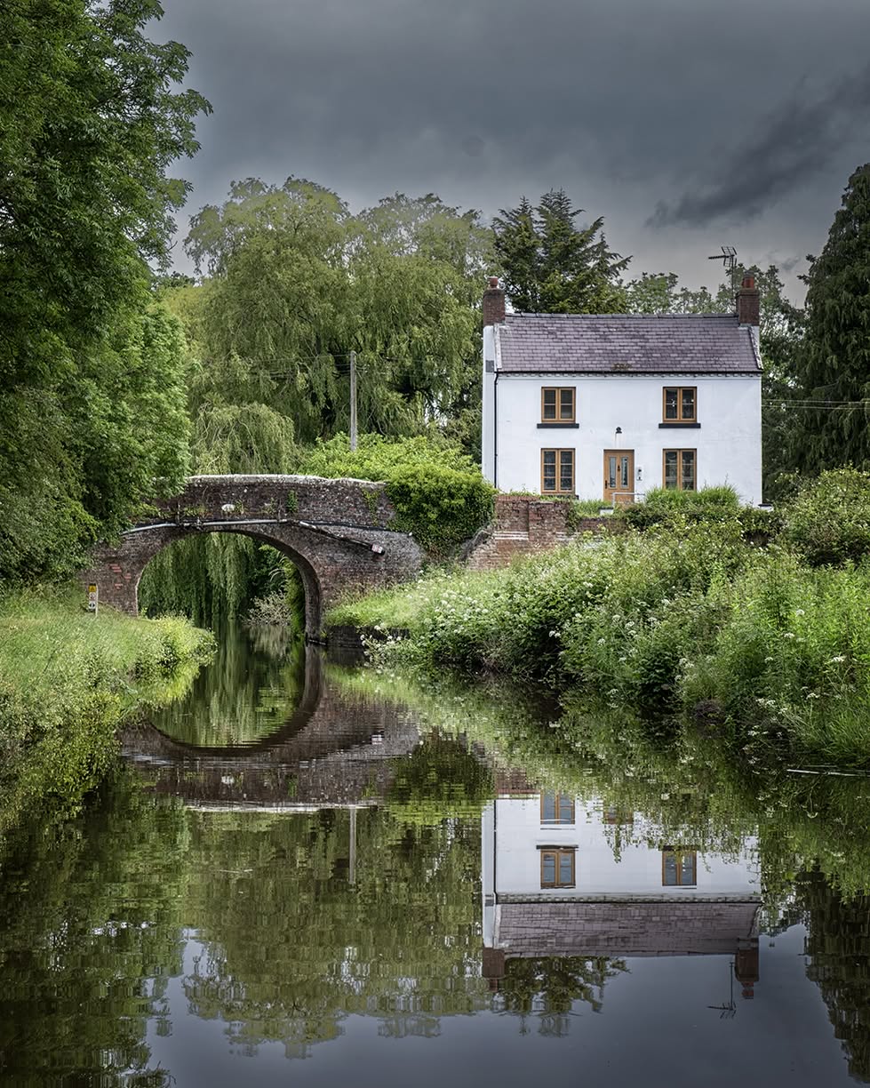 White house and stone bridge reflected in a calm canal surrounded by trees and greenery