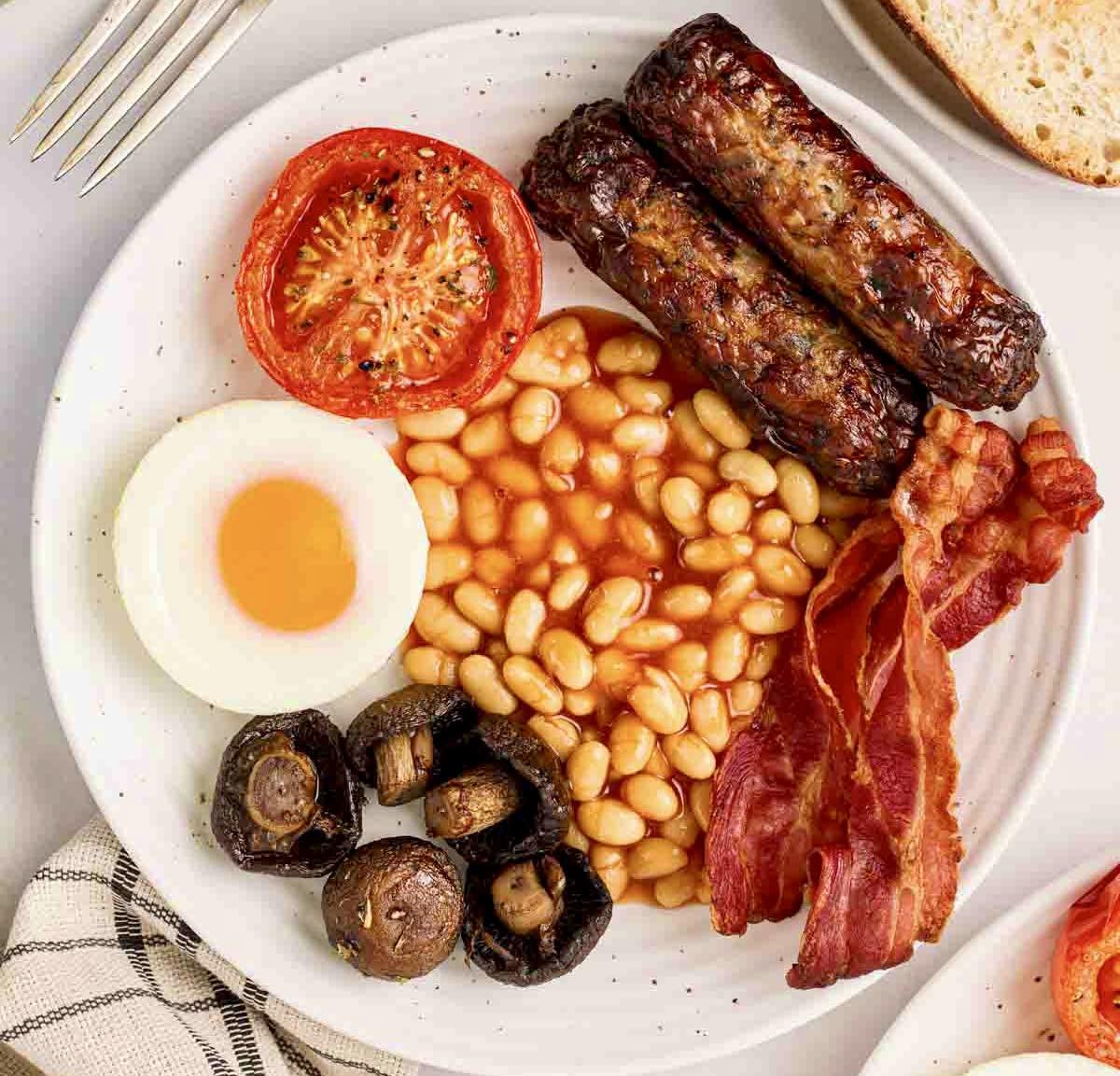 Full English breakfast with sausages, bacon, egg, beans, mushrooms and tomato on white plate