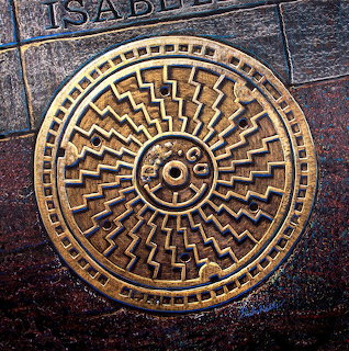 Decorative bronze manhole cover with concentric geometric patterns on dark stone pavement