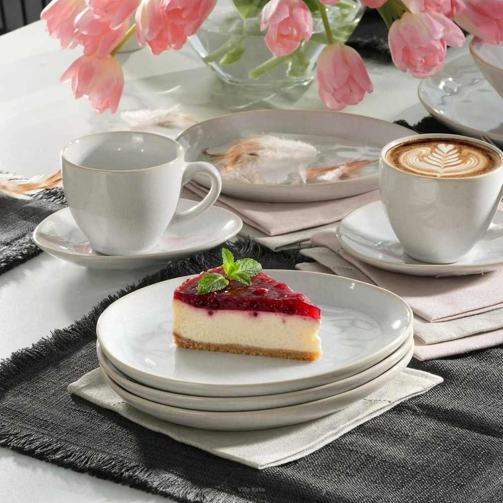 Slice of cheesecake with red berry topping on white plate next to two coffee cups and pink tulips