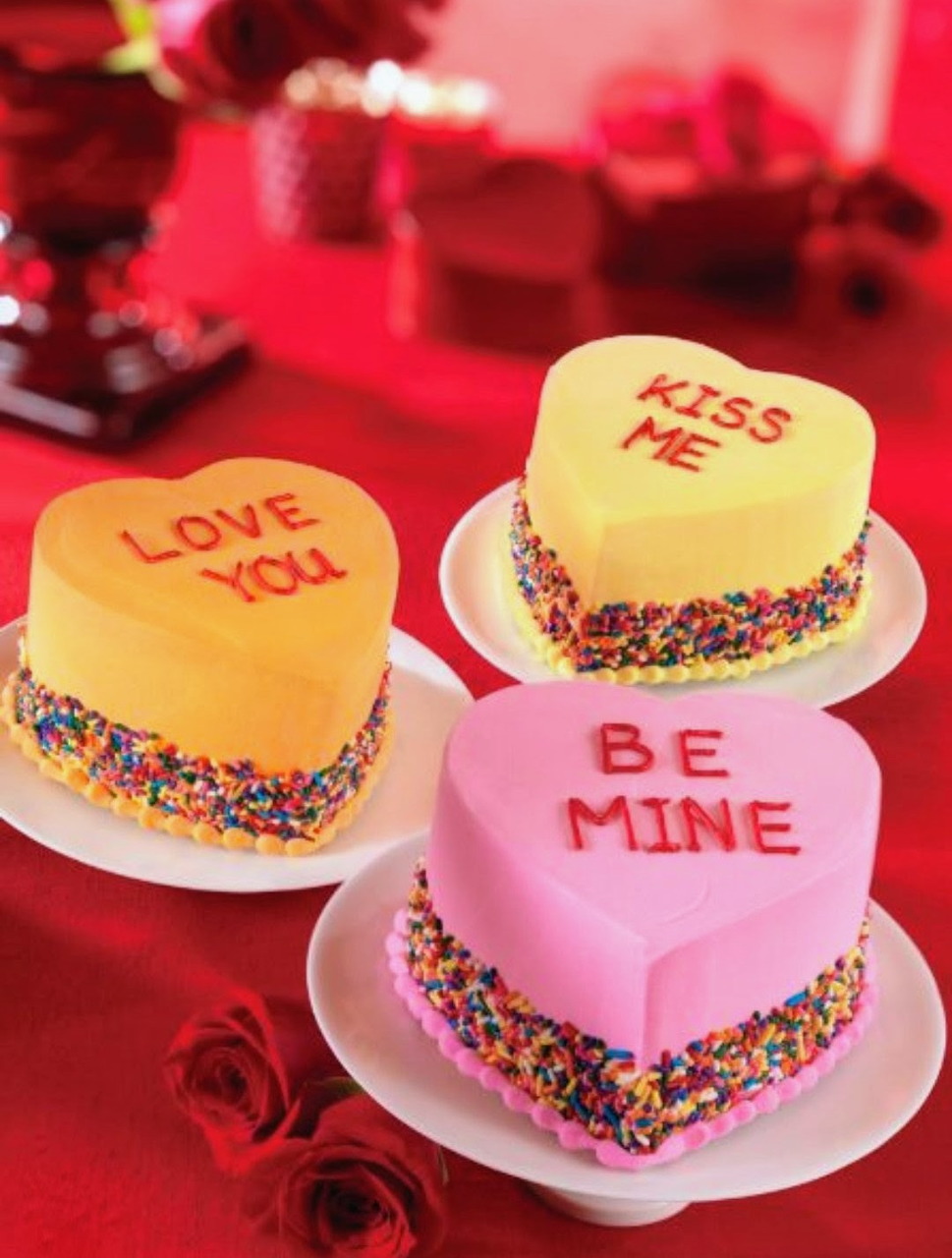 Three heart-shaped cakes with Valentine messages on white plates, decorated with sprinkles and surrounded by red roses