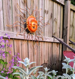 Decorative sun face on a wooden fence with purple flowers and silvery garden plants