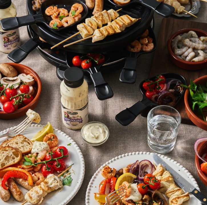 Overhead view of a raclette grill surrounded by shrimp skewers, chicken skewers, garlic sauce, and side dishes