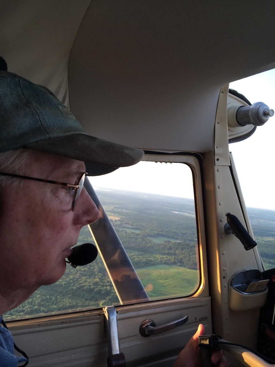 Man in cap and glasses flying a small plane with green landscape visible through cockpit window