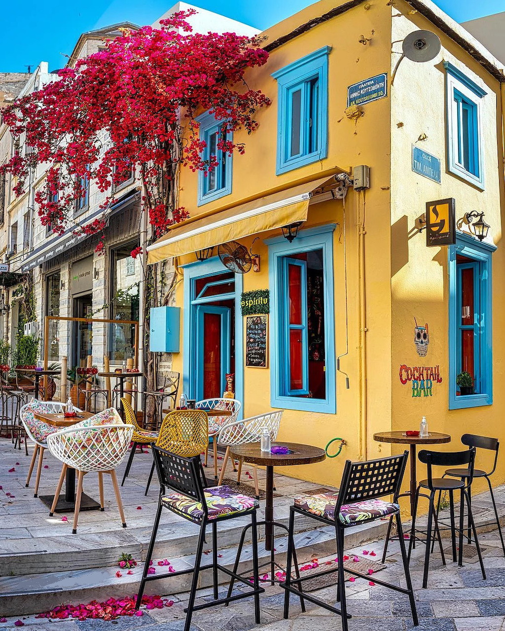 Yellow building with blue shutters, outdoor cafe seating, and bright pink flowering tree on Mediterranean street