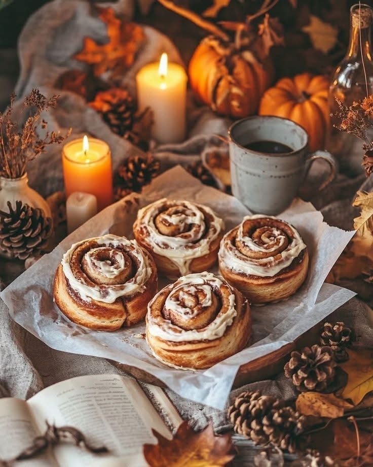 Cinnamon rolls with candles, pumpkins, coffee, pinecones, and an open book in a cozy autumn setting