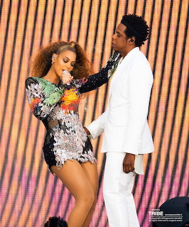 Two performers on stage, woman in sequined bodysuit and man in white suit with striped orange backdrop