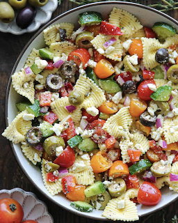 Bowl of pasta salad with farfalle, tomatoes, olives, feta cheese, avocado, and herbs on a wooden table