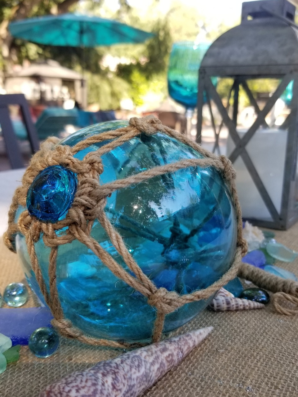 Blue glass fishing float with rope netting on burlap surface beside a metal lantern