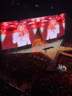 Indoor concert arena with star-shaped stage and large screens showing a female performer