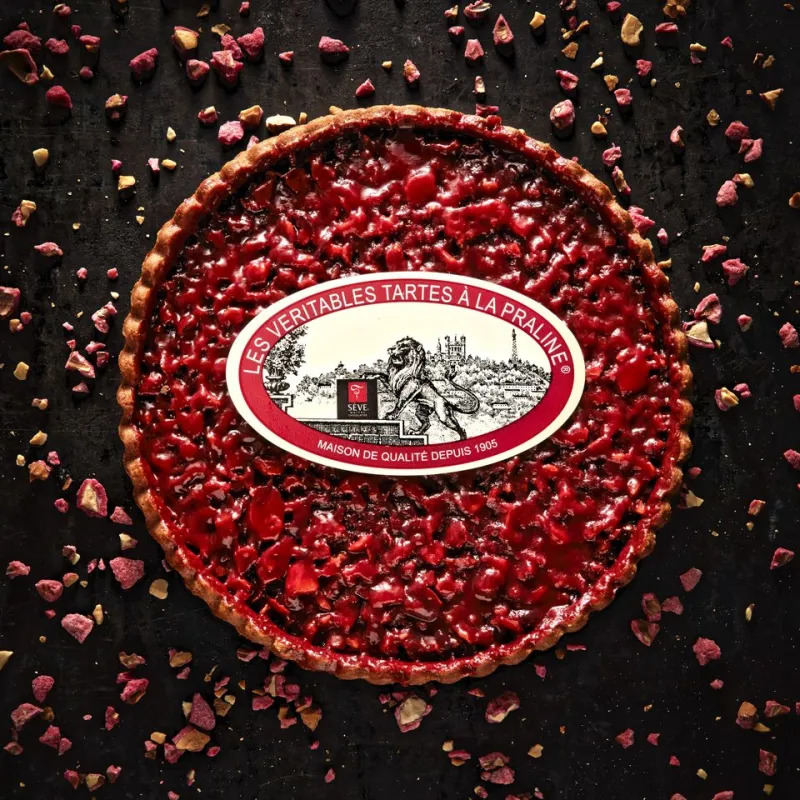 Round tart filled with pink pralines and decorative vintage label on dark background with scattered praline pieces