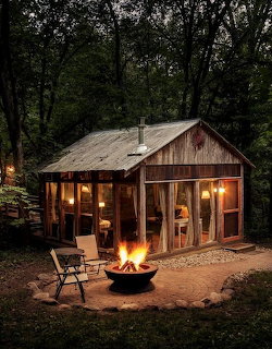 Rustic cabin in forest at dusk with warm interior lights and campfire in foreground