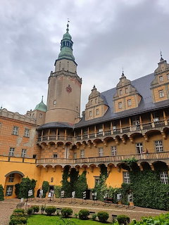 Renaissance palace courtyard with clock tower, arcaded galleries, and manicured garden under cloudy sky