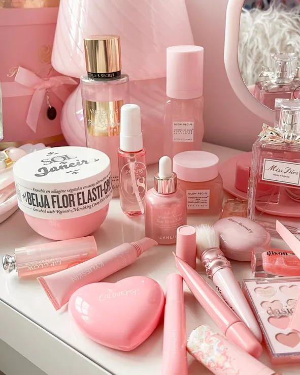 Pink cosmetic and beauty products displayed on a vanity table with perfume, lip gloss, cream jars, and tools