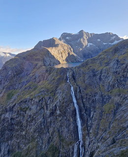 Tall waterfall streaming down rocky cliffs with a glacial lake and mountain peaks in New Zealand