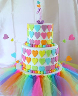 Pastel rainbow heart-decorated birthday cake with number one candle on colorful tulle base