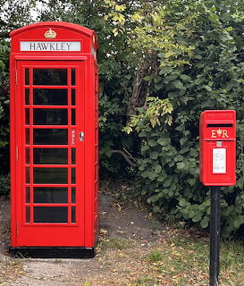Red British telephone box marked Hawkley next to a red Royal Mail post box on a leafy rural path in England