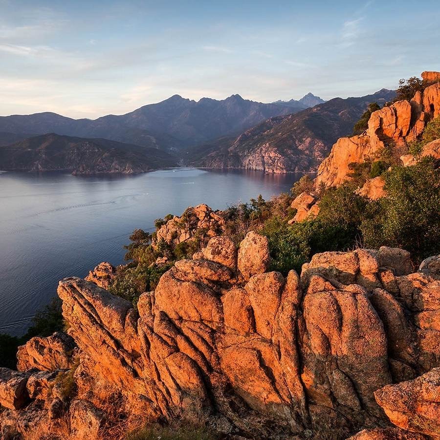 Red granite rock formations above a blue bay in Calanques de Piana, Corsica with mountain backdrop