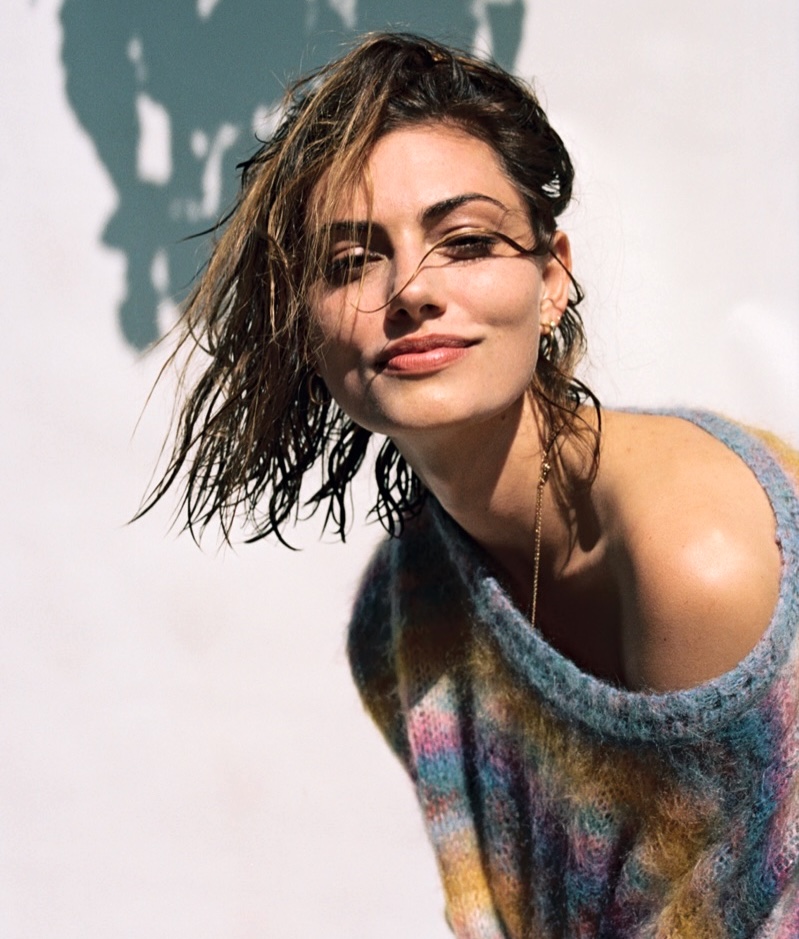 Woman with wet hair in a colorful knit sweater smiling in sunlight against a white wall
