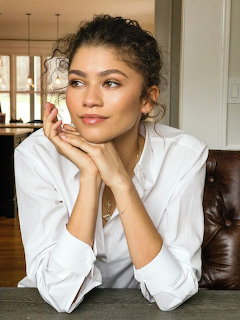 Young woman with curly updo hairstyle wearing white shirt, chin resting on hands, indoor setting
